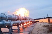A series of large metal pipes are positioned on a sandy surface, extending into the distance. There is a significant flame erupting from the end of the pipes, indicating a possible gas or oil pipeline. The sky is overcast with a grayish-blue hue and the area seems to be a remote, undeveloped area with some vegetation.