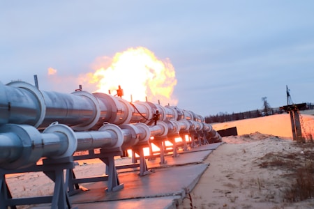 A series of large metal pipes are positioned on a sandy surface, extending into the distance. There is a significant flame erupting from the end of the pipes, indicating a possible gas or oil pipeline. The sky is overcast with a grayish-blue hue and the area seems to be a remote, undeveloped area with some vegetation.