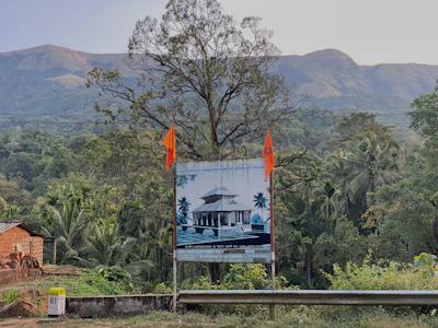 A scenic landscape featuring lush green trees and foliage with hills in the background. A large billboard is prominently displayed in the foreground, showing an image of a building and flanked by two orange flags. The scene includes a small structure to the left and a milestone marker in the bottom left corner, indicating distance.
