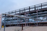 A complex network of large, industrial pipes is supported by a metal framework, situated on a sandy surface. The pipes are metallic and exhibit some signs of rust and wear. Overcast skies provide a muted backdrop to this industrial scene.