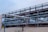 A complex network of large, industrial pipes is supported by a metal framework, situated on a sandy surface. The pipes are metallic and exhibit some signs of rust and wear. Overcast skies provide a muted backdrop to this industrial scene.