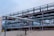 A complex network of large, industrial pipes is supported by a metal framework, situated on a sandy surface. The pipes are metallic and exhibit some signs of rust and wear. Overcast skies provide a muted backdrop to this industrial scene.