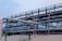 A complex network of large, industrial pipes is supported by a metal framework, situated on a sandy surface. The pipes are metallic and exhibit some signs of rust and wear. Overcast skies provide a muted backdrop to this industrial scene.