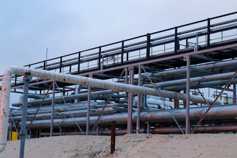A complex network of large, industrial pipes is supported by a metal framework, situated on a sandy surface. The pipes are metallic and exhibit some signs of rust and wear. Overcast skies provide a muted backdrop to this industrial scene.