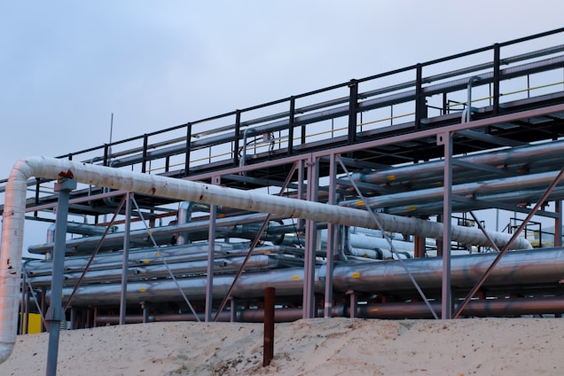 A complex network of large, industrial pipes is supported by a metal framework, situated on a sandy surface. The pipes are metallic and exhibit some signs of rust and wear. Overcast skies provide a muted backdrop to this industrial scene.