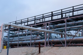 A complex network of large, industrial pipes is supported by a metal framework, situated on a sandy surface. The pipes are metallic and exhibit some signs of rust and wear. Overcast skies provide a muted backdrop to this industrial scene.