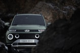 A green SUV with a modern design is positioned amidst rocky terrain. The vehicle's headlights are on, and droplets of water or snow are visible, indicating a damp or cold environment. The background is dark with blurred rocks framing the vehicle.
