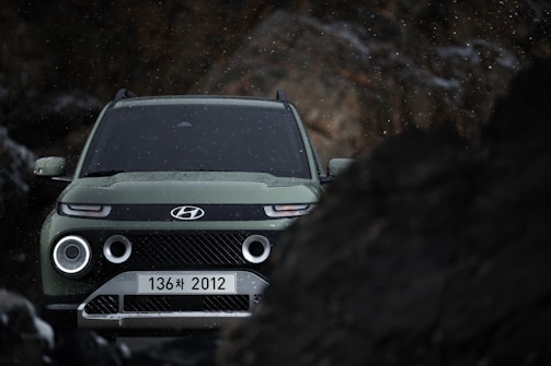 A green SUV with a modern design is positioned amidst rocky terrain. The vehicle's headlights are on, and droplets of water or snow are visible, indicating a damp or cold environment. The background is dark with blurred rocks framing the vehicle.