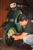 A person cleaning the front wheel of a dark green vintage scooter with a green cloth. The scooter is parked indoors against a wooden wall.