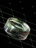 A transparent, reflective oval object with a textured surface is centered against a dark background. The object displays varied colors like green and blue, creating a prismatic effect. The background features a grid pattern of thin diagonal lines with small white dots at intersections, resembling a starry sky or futuristic design.