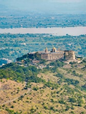 A historic fort situated on a hilltop surrounded by lush greenery and a sprawling landscape. The structure features traditional architecture with visible fort walls and towers. The foreground includes a winding path leading up to the fort. In the distance, a river and a vast area of flatlands can be seen, stretching to the horizon.
