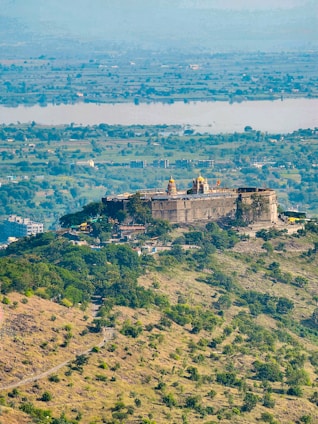 A historic fort situated on a hilltop surrounded by lush greenery and a sprawling landscape. The structure features traditional architecture with visible fort walls and towers. The foreground includes a winding path leading up to the fort. In the distance, a river and a vast area of flatlands can be seen, stretching to the horizon.