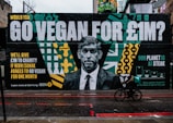 A large street mural features a grayscale portrait of a man in a suit. Beside the portrait are bold letters with a message about going vegan for a charity incentive of £1 million. The artwork includes vibrant patterns in green, yellow, and teal, along with graphs and text. A cyclist on a black bicycle is riding past on a wet road.