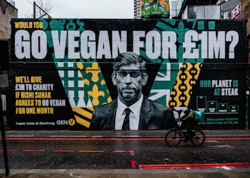 A large street mural features a grayscale portrait of a man in a suit. Beside the portrait are bold letters with a message about going vegan for a charity incentive of &pound;1 million. The artwork includes vibrant patterns in green, yellow, and teal, along with graphs and text. A cyclist on a black bicycle is riding past on a wet road.