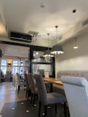 A cozy, modern restaurant interior featuring wooden tables and upholstered chairs in grey tones. The room is well-lit with hanging lamps and natural light streaming through large windows. The decor includes a stylish partition with some decorative items on shelves and a mix of seating arrangements with both individual and long bench-style options.