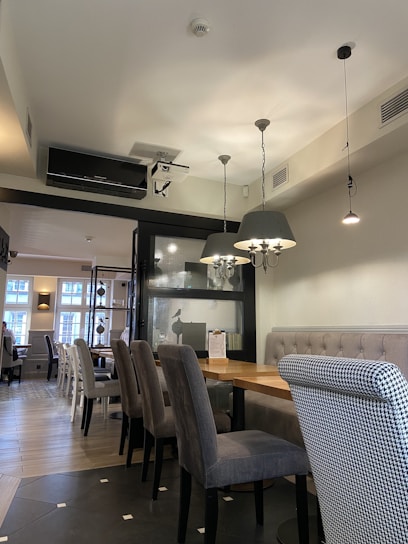 A cozy, modern restaurant interior featuring wooden tables and upholstered chairs in grey tones. The room is well-lit with hanging lamps and natural light streaming through large windows. The decor includes a stylish partition with some decorative items on shelves and a mix of seating arrangements with both individual and long bench-style options.