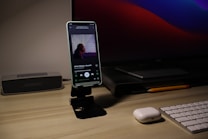 A smartphone displaying a music player app is placed on a sleek stand on a wooden desk. Next to it, there is a wireless speaker on the left and a modern computer monitor in the background. In front of the monitor, there is a closed laptop and a pencil resting beside it. An AirPods case is positioned near a white wireless keyboard on the right side.