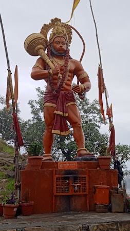 A large statue of a deity with red-orange skin, holding a mace and wearing a golden crown, a garland, and a red garment. The statue stands on a decorated platform surrounded by plants and flags waving in the air. Behind it are lush green trees, and the sky appears overcast.