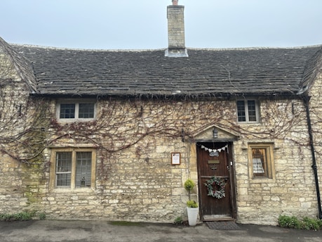 A rustic stone cottage with a tiled roof and a prominent chimney centered at the top. The facade is adorned with climbing vines and features three windows with metal grilles. A wooden door is decorated with a wreath and a 'closed' sign. Two potted plants flank the entrance.