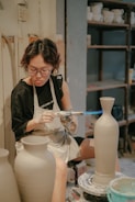 A person is using a blowtorch to work on a ceramic vase in a pottery studio. They are wearing an apron and glasses, focused intently on their craft. Shelves filled with various pottery items are visible in the background.