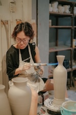 A person is using a blowtorch to work on a ceramic vase in a pottery studio. They are wearing an apron and glasses, focused intently on their craft. Shelves filled with various pottery items are visible in the background.