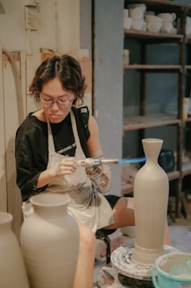A person is using a blowtorch to work on a ceramic vase in a pottery studio. They are wearing an apron and glasses, focused intently on their craft. Shelves filled with various pottery items are visible in the background.