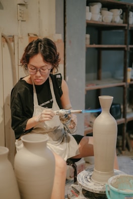 A person is using a blowtorch to work on a ceramic vase in a pottery studio. They are wearing an apron and glasses, focused intently on their craft. Shelves filled with various pottery items are visible in the background.