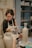 A person is using a blowtorch to work on a ceramic vase in a pottery studio. They are wearing an apron and glasses, focused intently on their craft. Shelves filled with various pottery items are visible in the background.