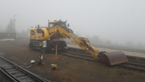 A yellow construction excavator is positioned on a railway track amidst a foggy environment. The equipment, labeled with 'Liebherr,' has its arm extended downwards with a metal scoop attachment. The surrounding area features a misty background with limited visibility, some gravel, and railway tracks.