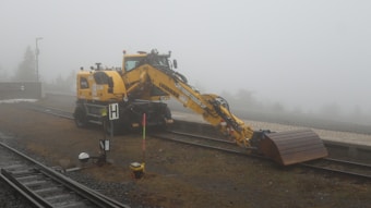 A yellow construction excavator is positioned on a railway track amidst a foggy environment. The equipment, labeled with 'Liebherr,' has its arm extended downwards with a metal scoop attachment. The surrounding area features a misty background with limited visibility, some gravel, and railway tracks.