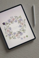 An iPad displays a digital illustration of delicate flowers arranged in a wreath-like pattern. The floral design consists of soft purple and pink petals with muted green leaves. Next to the iPad is a white stylus, resting on a textured light gray surface.