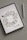 An iPad displays a digital illustration of delicate flowers arranged in a wreath-like pattern. The floral design consists of soft purple and pink petals with muted green leaves. Next to the iPad is a white stylus, resting on a textured light gray surface.