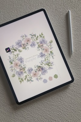 An iPad displays a digital illustration of delicate flowers arranged in a wreath-like pattern. The floral design consists of soft purple and pink petals with muted green leaves. Next to the iPad is a white stylus, resting on a textured light gray surface.