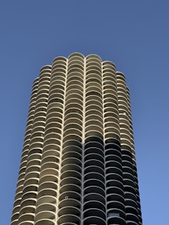 A tall cylindrical building with a repetitive pattern of curved balconies extending upwards against a clear blue sky. The architectural design features symmetrical, stacked disk-like floors, creating a modern and sleek appearance.