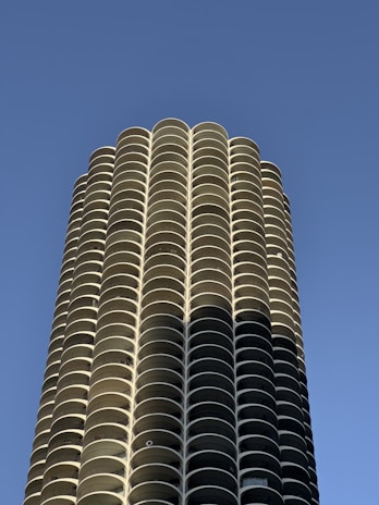 A tall cylindrical building with a repetitive pattern of curved balconies extending upwards against a clear blue sky. The architectural design features symmetrical, stacked disk-like floors, creating a modern and sleek appearance.