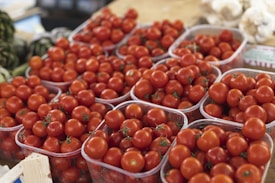 A collection of ripe red tomatoes neatly arranged in plastic containers, possibly displayed at a market or grocery setting. The tomatoes are vibrant and appear fresh, with visible green stems. In the background, there are hints of other vegetables like artichokes and garlic.