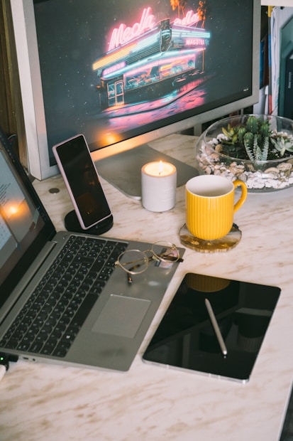 A cozy workspace setup featuring a laptop, tablet with a stylus, smartphone, a lit candle, eyeglasses, and a yellow mug on a marble desk. A monitor in the background displays a neon-lit diner scene. A bowl of small cacti and pebbles adds a touch of greenery.