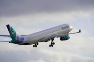 An airplane with turquoise and purple accents is ascending into the cloudy sky. The aircraft is primarily white with the word 'Iberojet' visible on the side. The landing gear is in the process of being retracted.