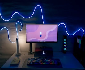 A modern desk setup featuring a widescreen monitor displaying a minimalistic pink and white abstract background. A colorful RGB keyboard and a mouse sit on the white desk, which also holds a laptop with a glowing logo. Above, blue LED strip lights create a wavy pattern on the wall, adding a futuristic ambiance. A white, sleek desk lamp is also present beside a book titled 'Do Epic Shit'.