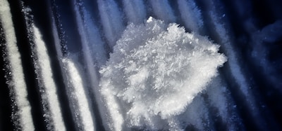 An intricate cluster of ice crystals sits atop a dark, ribbed surface, with detailed snowflake structures visible.
