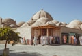A traditional marketplace with colorful clothing displayed on racks outside a brick building. The background features several domed structures, commonly found in Middle Eastern architecture. A person is casually walking by, browsing the garments under a bright, clear sky. A small tree with green leaves is in the foreground, adding a touch of nature to the scene.