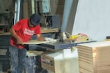 A person wearing a red T-shirt and a dark cap is working in a woodworking shop. The individual is using a machine to process a piece of wood. The shop is filled with various woodworking tools, equipment, and supplies. There is a stack of wood on the right side of the image.