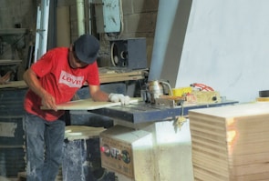 A person wearing a red T-shirt and a dark cap is working in a woodworking shop. The individual is using a machine to process a piece of wood. The shop is filled with various woodworking tools, equipment, and supplies. There is a stack of wood on the right side of the image.