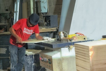 A person wearing a red T-shirt and a dark cap is working in a woodworking shop. The individual is using a machine to process a piece of wood. The shop is filled with various woodworking tools, equipment, and supplies. There is a stack of wood on the right side of the image.