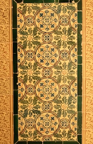 A series of ornate ceramic tiles featuring intricate floral and geometric patterns. The primary design consists of interlocking circles and stylized leaves in blue and green hues, set against a cream background. The border is formed by dark green tiles, and an adjacent area has a more delicate floral motif in brown and cream tones.