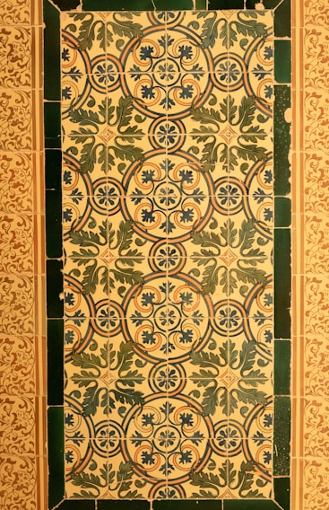 A series of ornate ceramic tiles featuring intricate floral and geometric patterns. The primary design consists of interlocking circles and stylized leaves in blue and green hues, set against a cream background. The border is formed by dark green tiles, and an adjacent area has a more delicate floral motif in brown and cream tones.