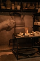 A pottery workshop with shelves displaying various clay pots and vases. A potter's hands are captured in motion while shaping clay on a wheel. A table in the foreground holds several unfinished clay pieces and pottery tools, illuminated by warm lighting.