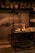 A pottery workshop with shelves displaying various clay pots and vases. A potter's hands are captured in motion while shaping clay on a wheel. A table in the foreground holds several unfinished clay pieces and pottery tools, illuminated by warm lighting.