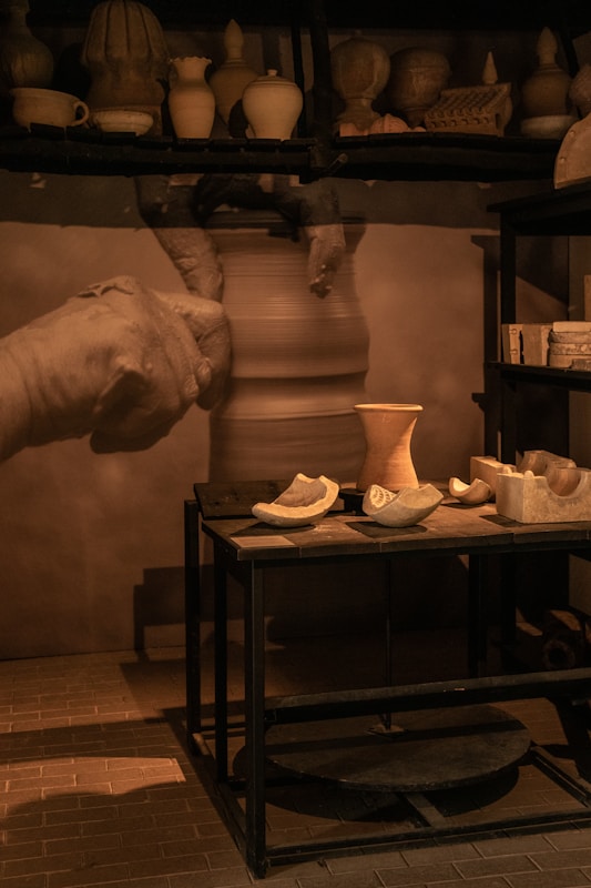 A pottery workshop with shelves displaying various clay pots and vases. A potter's hands are captured in motion while shaping clay on a wheel. A table in the foreground holds several unfinished clay pieces and pottery tools, illuminated by warm lighting.