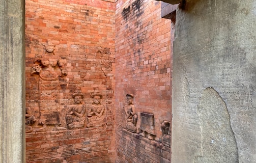 The image features ancient brick walls adorned with detailed bas-relief sculptures depicting figures in traditional attire. On the right, an engraved stone tablet includes several lines of intricate script, possibly an ancient language or inscription.